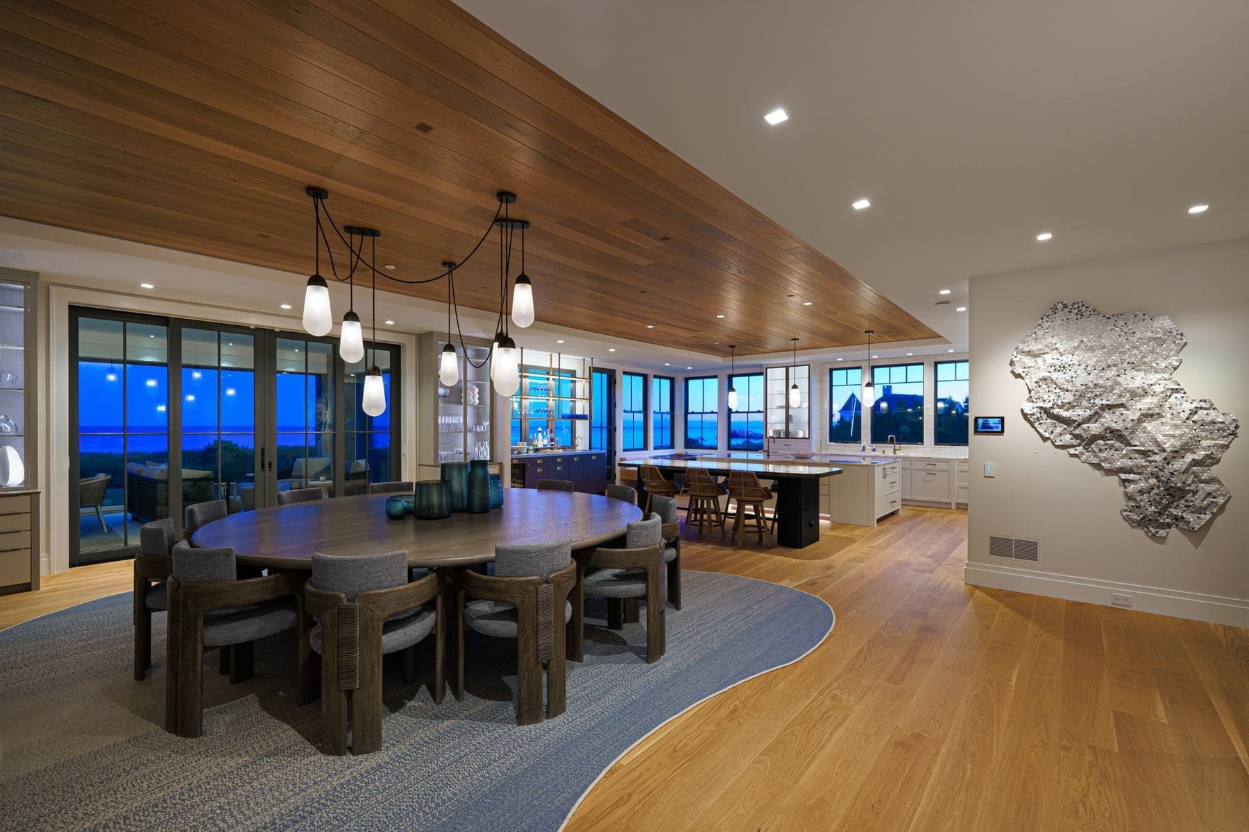 Open dining room and kitchen at dusk with ocean views through floor-to-ceiling windows and warm architectural lighting